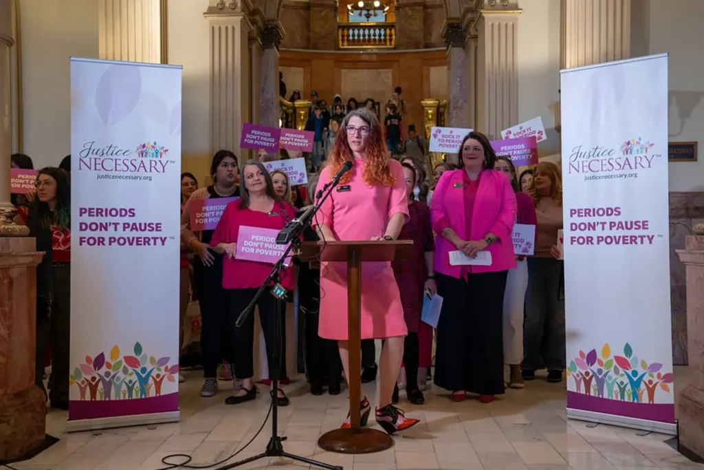 A Justice Necessary representative speaking at a podium, with other volunteers behind her holding "Periods don't pause for poverty" signs in a government building lobby.