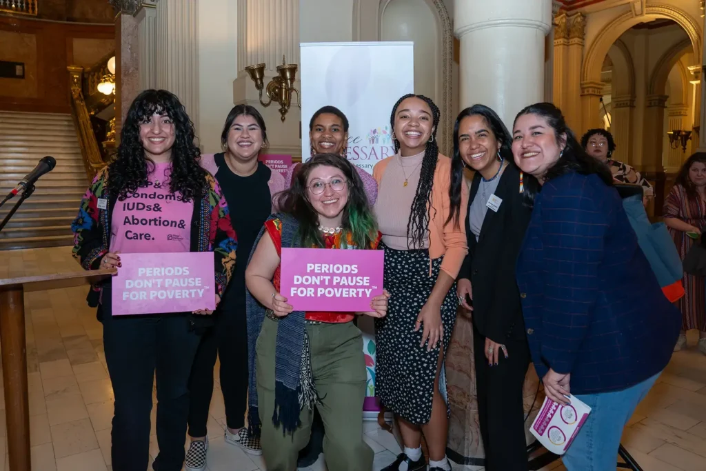 A group of ladies posing in the lobby of a government building, holding signs that say "Periods Don't Pause for Poverty"