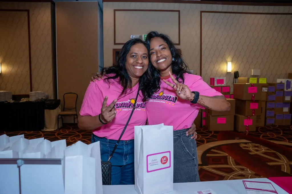 Two women wearing PeriodPalooza shirts wrap their arms around each other and raise peace signs with their hands